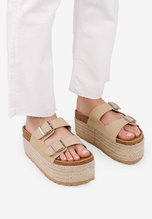 Sandalias de plataforma beige con dos correas ajustables, suela tejida estilo yute y base de goma texturizada. El modelo lleva pantalones cortos blancos.