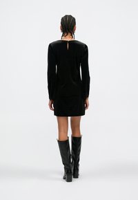 Vestido preto de veludo com mangas compridas e design de costas com abertura em gota, combinado com botas de couro pretas até ao joelho e textura suave.