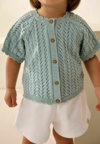 Hellblauer gestrickter Cardigan mit kurzen Ärmeln, der ein strukturiertes Muster aufweist und fünf Holzknöpfe auf der Vorderseite hat. Über weißen Shorts getragen.