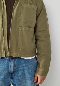 Olive green zip-up jacket with two chest pockets, made of sturdy fabric. Paired with light blue denim jeans, displaying a relaxed fit.