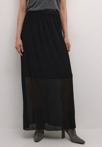 Schwarzer Maxikleid mit fließendem Design, ausgestattet mit transparenten Einsätzen am unteren Ende und einem elastischen Bund. Kombiniert mit metallischen High-Heel-Stiefeln.