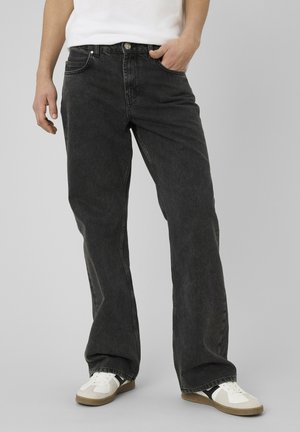 Personne portant un jean noir délavé ample et des baskets blanches, une main dans la poche, sur un fond gris uni.