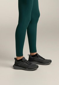 Czarne buty sportowe z teksturowaną cholewką, sznurowanym zapięciem i amortyzowaną podeszwą, uzupełnione zielonymi dopasowanymi legginsami.