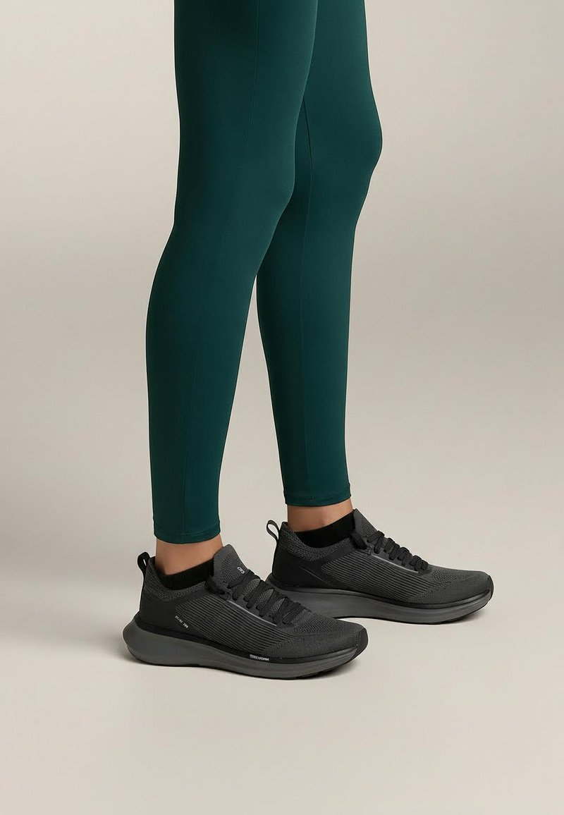 Czarne buty sportowe z teksturowaną cholewką, sznurowanym zapięciem i amortyzowaną podeszwą, uzupełnione zielonymi dopasowanymi legginsami.