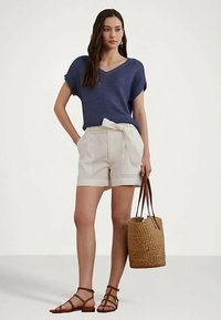 Γυναίκα με μπλε κοντομάνικη πλεκτή μπλούζα, λευκά σορτς ψηλής μέσης με ζώνη, καφέ σανδάλια με λουριά, κρατώντας μια υφασμάτινη τσάντα.