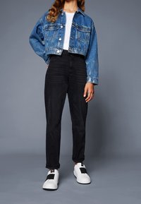 Ořezaná modrá denimová bunda se dvěma předními kapsami, spárovaná s černými džíny s vysokým pasem a bílými slip-on teniskami s černými akcenty.