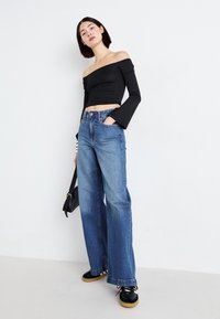 Black off-the-shoulder top with long sleeves, paired with wide-leg blue denim jeans and black sneakers with gum soles. Black bag details visible.