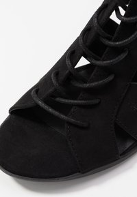 Czarne zamszowe sandały z otwartym noskiem, z krzyżującymi się sznurkami i wycięciami. Płaska podeszwa i gładka wewnętrzna tekstura.