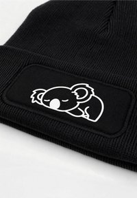 Black knit beanie featuring a textured ribbed pattern and a white patch with an illustration of a sleeping koala.