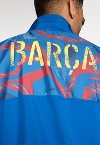 Modrá sportovní bunda s texturovanou tkaninou v červených a šedých vzorech. "BARÇA" v tučných, opotřebovaných žlutých písmenech na horní části zad.