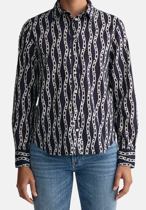 Granatowa koszula na guziki z białym wzorem łańcucha, noszona z niebieskimi dżinsami, pokazana od szyi do bioder na jednolitym tle.