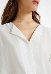 Primo piano del décolleté e delle labbra di una donna, che indossa una blusa bianca con colletto a V, con lunghi capelli castani in vista.