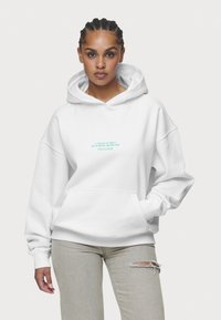 Bílá mikina s kapsou ve stylu klokan, ve středu s tyrkysovým textem. Spolu s světle šedými džíny s opotřebovanými detaily.