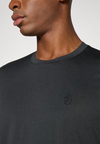 Svart ribbestrikket t-skjorte med rund hals, prydet med en diskret brodert logo på brystet. Stoffet har en glatt, teksturert overflate.