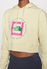 Hoodie beige cropped con logo grafico verde e rosa; dotata di cappuccio con cordino e orlo a costine. Tessuto morbido con una texture liscia.