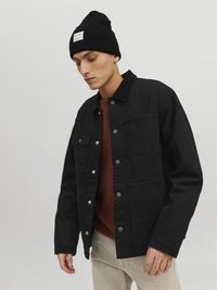 Chaqueta de mezcla de algodón negra con botones a presión, dos bolsillos en el pecho y un cuello de pana, llevada sobre un suéter de punto marrón y pantalones claros. Gorro negro en la cabeza.