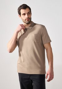 Beige poolopaita sileässä kankaassa, jossa on lyhyt kaulus, puoliveto ja rintatasku. Yhdistetty tummaan housuun, malli säätää kaulusta.