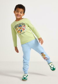 Grünes Langarmhemd mit Cartoon-Hunden, kombiniert mit hellblauen Jogginghosen mit Charakterdesigns und grünen Sneakers mit weißen Akzenten.