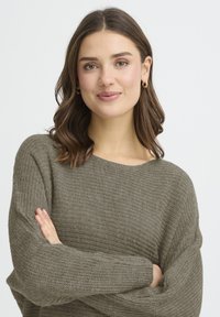 Pull tricot vert kaki à coupe ample, texture côtelée et large encolure. Le mannequin pose les bras croisés, mettant en valeur le design.