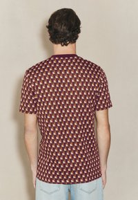 Tričková košeľa s krátkym rukávom so vzorom zloženým z trojuholníkov v bordovej, béžovej a krémovej farbe. Vyrobená z mäkkej látky s okrúhlym výstrihom.