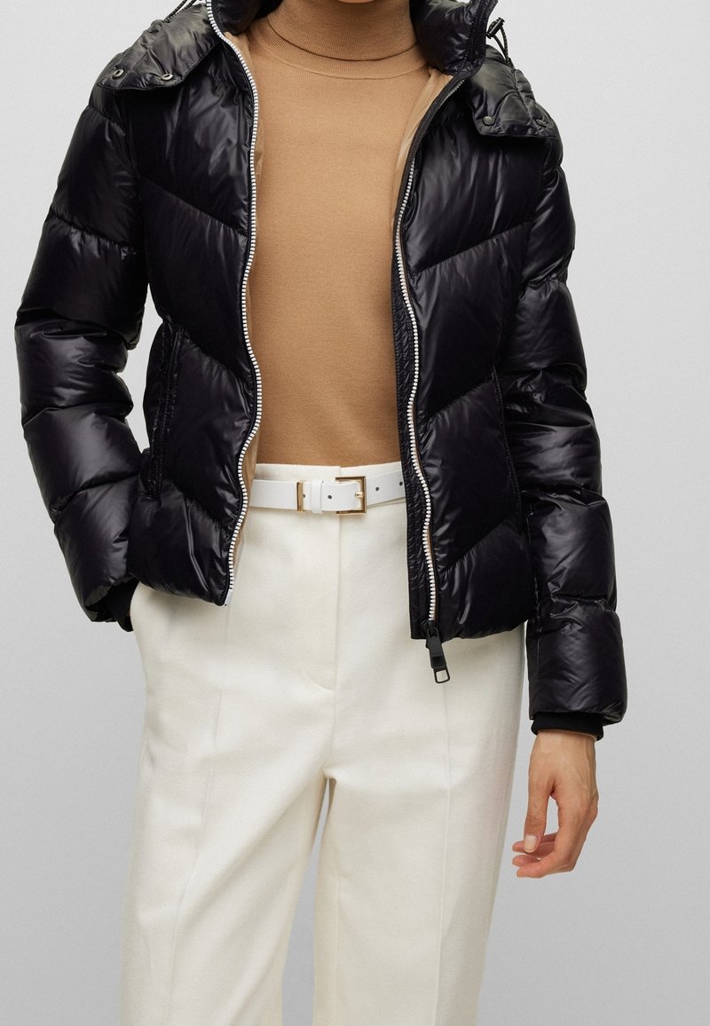 Péřová bunda, lesklý černý materiál, zipové zapínání, kapuce, přiléhavý střih. Nositá přes krémový rolák, doplněná bílými kalhotami s vysokým pasem.