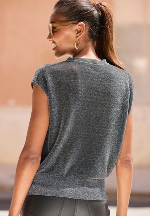 Frau mit braunen Haaren im Pferdeschwanz, die eine Sonnenbrille, goldene Ohrringe, ein ärmelloses silbernes Oberteil und schwarze Hosen trägt, schaut weg und lächelt.