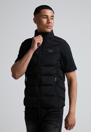 Jeune homme portant une doudoune noire sans manches sur un t-shirt noir à manches courtes, en train de fermer la doudoune, debout devant un fond gris uni.