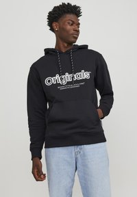 Sudadera con capucha negra y logo blanco "originals", detalles con cordones y un bolsillo frontal. Combinada con jeans de color azul claro. Corte relajado.