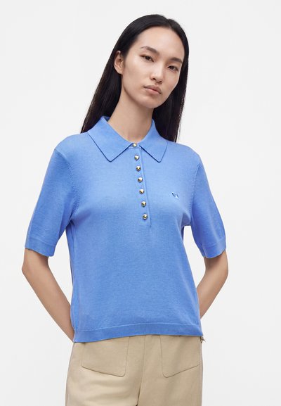 Femme portant un polo bleu clair à manches courtes avec des boutons dorés et un pantalon beige, debout devant un fond blanc uni.