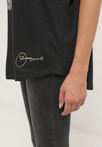 Černá košile s krátkým rukávem a zakřiveným lemem, na dolním levém rohu s vyšitým zlatým logem. Měkká, strukturovaná tkanina.