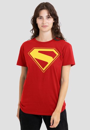 Woman with wavy brown hair wearing a red t-shirt with a large yellow geometric "S" logo, standing against a plain light gray background.