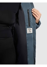 Lengva mėlyna pūsta striukė su užtrauktuku užsegamu kišene ir audinio etikete su užrašu „Šiltas apkabinimas iš audinio“. Vidus yra lygaus audinio.