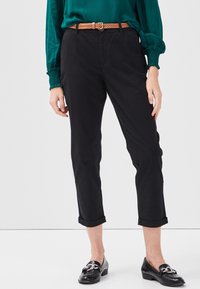 Personne portant un pantalon noir court, un chemisier vert à manches longues, une ceinture tressée beige et des mocassins noirs avec un détail en chaîne argentée.