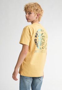 Gul bomulds t-shirt med korte ærmer, der har et flerfarvet grafik af palmeblade og tekst på bagsiden samt et mærke ved kanten.