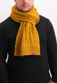 Knitted scarf in mustard yellow, featuring a chunky cable pattern, ribbed ends, and a brown leather label, worn over a black sweater.