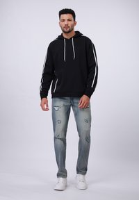 Zwarte hoodie met witte zijstrepen; gecombineerd met versleten blauwe spijkerbroeken. Het model draagt witte sneakers en staat tegen een lichte achtergrond.