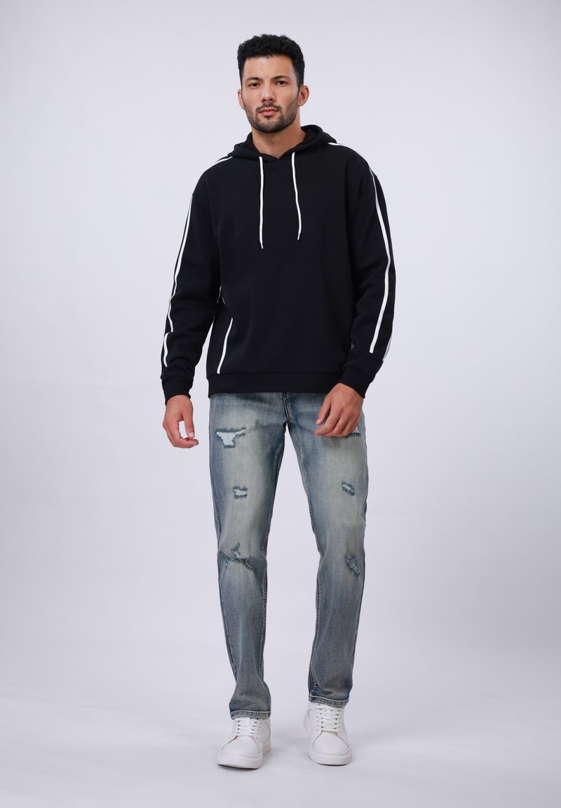 Felpa nera con cappuccio e strisce bianche sui lati; abbinata a jeans blu strappati. Il modello indossa sneakers bianche, in piedi contro uno sfondo chiaro.