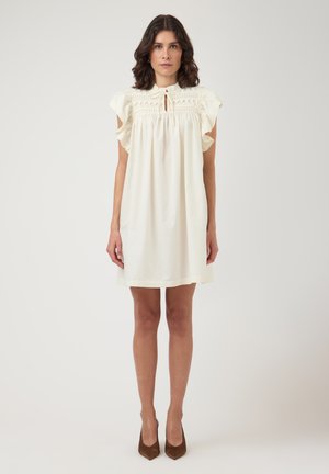 Woman standing facing forward wearing a knee-length cream dress with ruffled sleeves and brown pointed heels against a plain background.