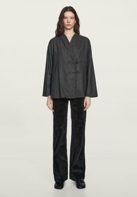 Sort tekstureret jakke med en wrap-design, der har en Mandarin-krave og to knapper. Sæt sammen med lige sorte velourbukser.