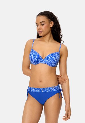 Mujer con bikini azul estampado, con tirantes ajustables y lazos laterales, de pie contra un fondo liso y claro.