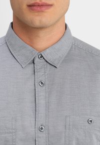 Chemise grise à boutons, dotée d'un col, de deux poches poitrine et de boutons contrastants. Confectionnée dans un tissu texturé.