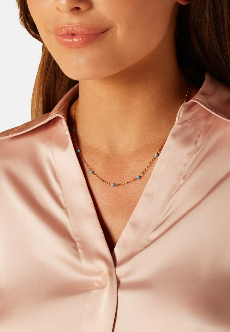 Gouden ketting met kleine turquoise kralen, gedragen over een lichtroze satijnen blouse. Gladde textuur en delicaat ontwerp.