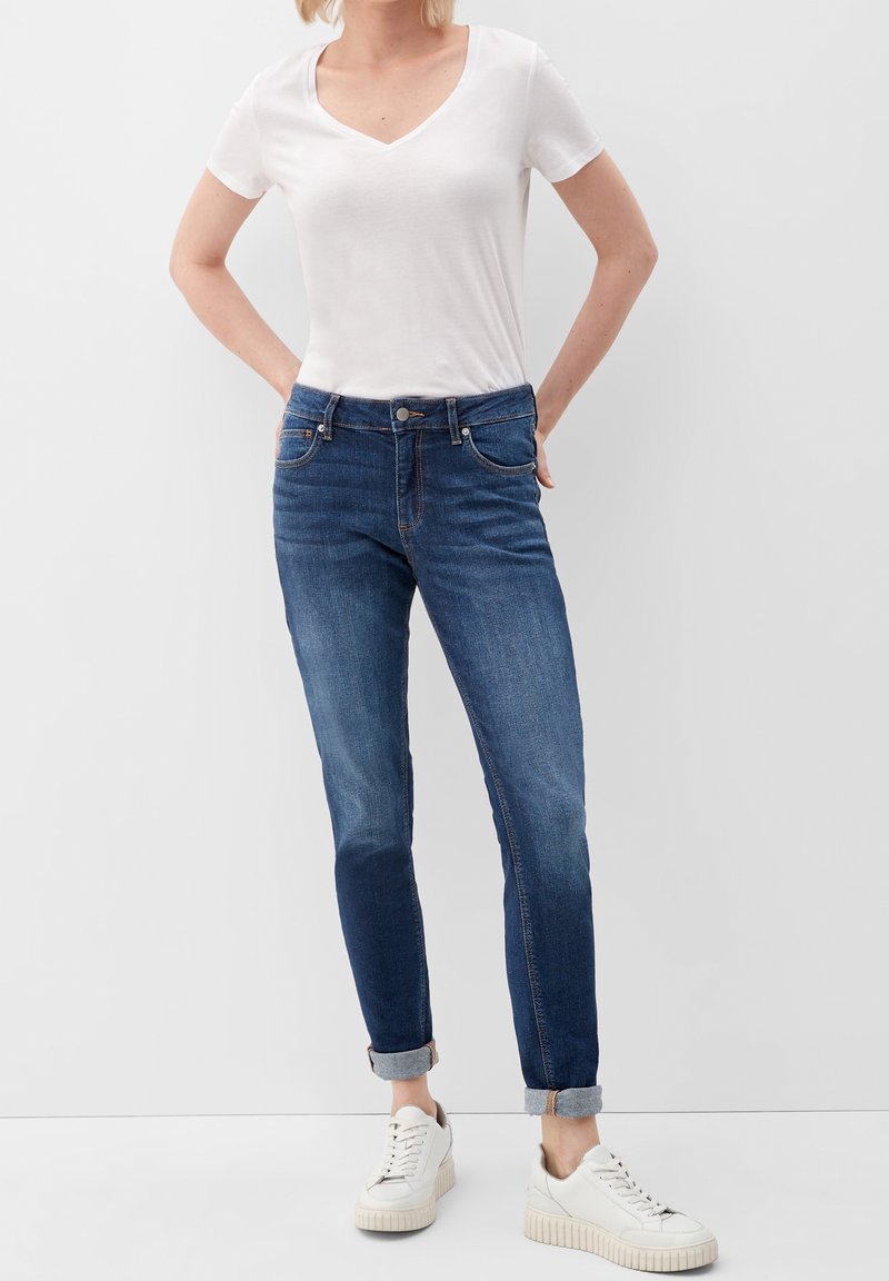 Camiseta blanca de cuello en V, vaqueros ajustados azules con dobladillos remangados y zapatillas blancas con suelas texturizadas. Los vaqueros tienen bolsillos delanteros y cierre de botón.