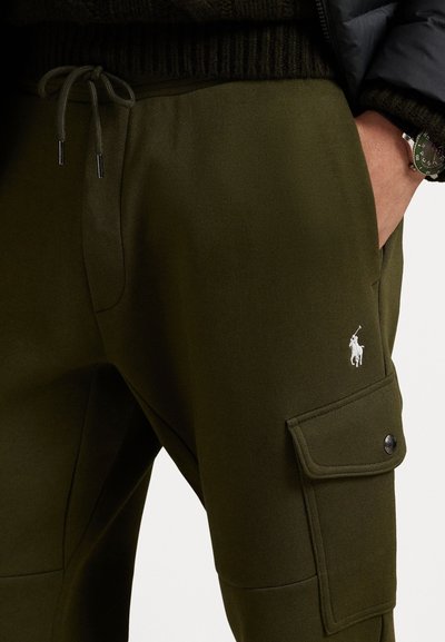 Kratki kadar maslinastozeleni trenirke s remenom, bočnim džepom za teret i bijelim logotipom Polo Ralph Lauren, te rukom u prednjem džepu.
