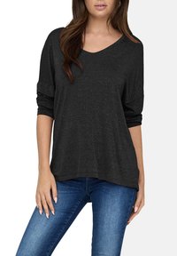 Top negro de cuello en V con mangas largas, hecho de una tela texturizada que presenta sutiles destellos. Corte holgado con un dobladillo curvado, combinado con jeans azules.