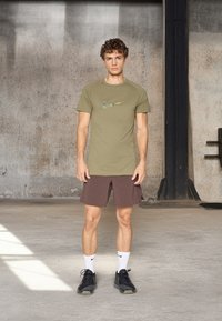 T-shirt atlético verde-oliva com um logótipo da Nike texturizado, combinado com calções castanhos e sapatilhas atléticas pretas, num espaço interior.