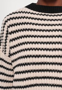 Gros plan d’un pull tricoté avec des rayures horizontales crème et noires et un col rond côtelé noir uni.