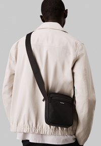 Borsa a tracolla nera con zip, realizzata in materiale testurizzato, indossata sopra una giacca beige chiaro con orlo elasticizzato, caratterizzata da un logo prominente.