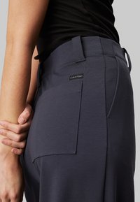 Modré elastické kalhoty s zadní kapsou a bočními smyčkami. Materiál má hladkou texturu s jemným leskem. Viditelné logo Calvin Klein.