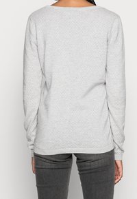 Pull en laine texturé gris clair avec un motif de diamant, des manches longues et une encolure large. Un jean gris coordonné est partiellement visible.